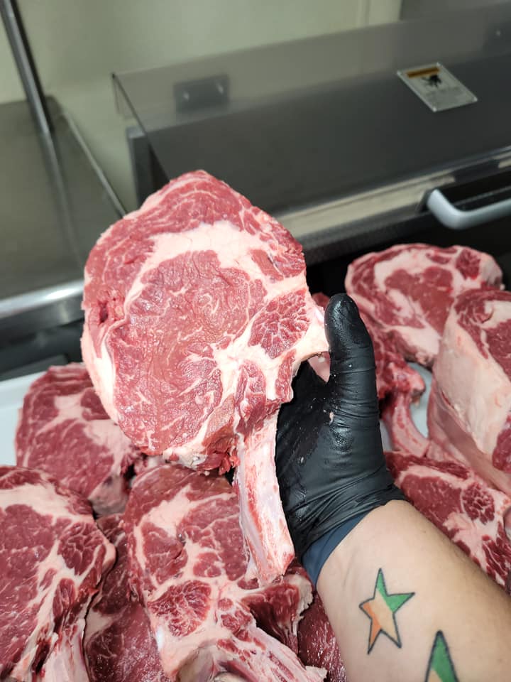 person with black glove on holding a raw tomahawk steak above other tomahawk steaks